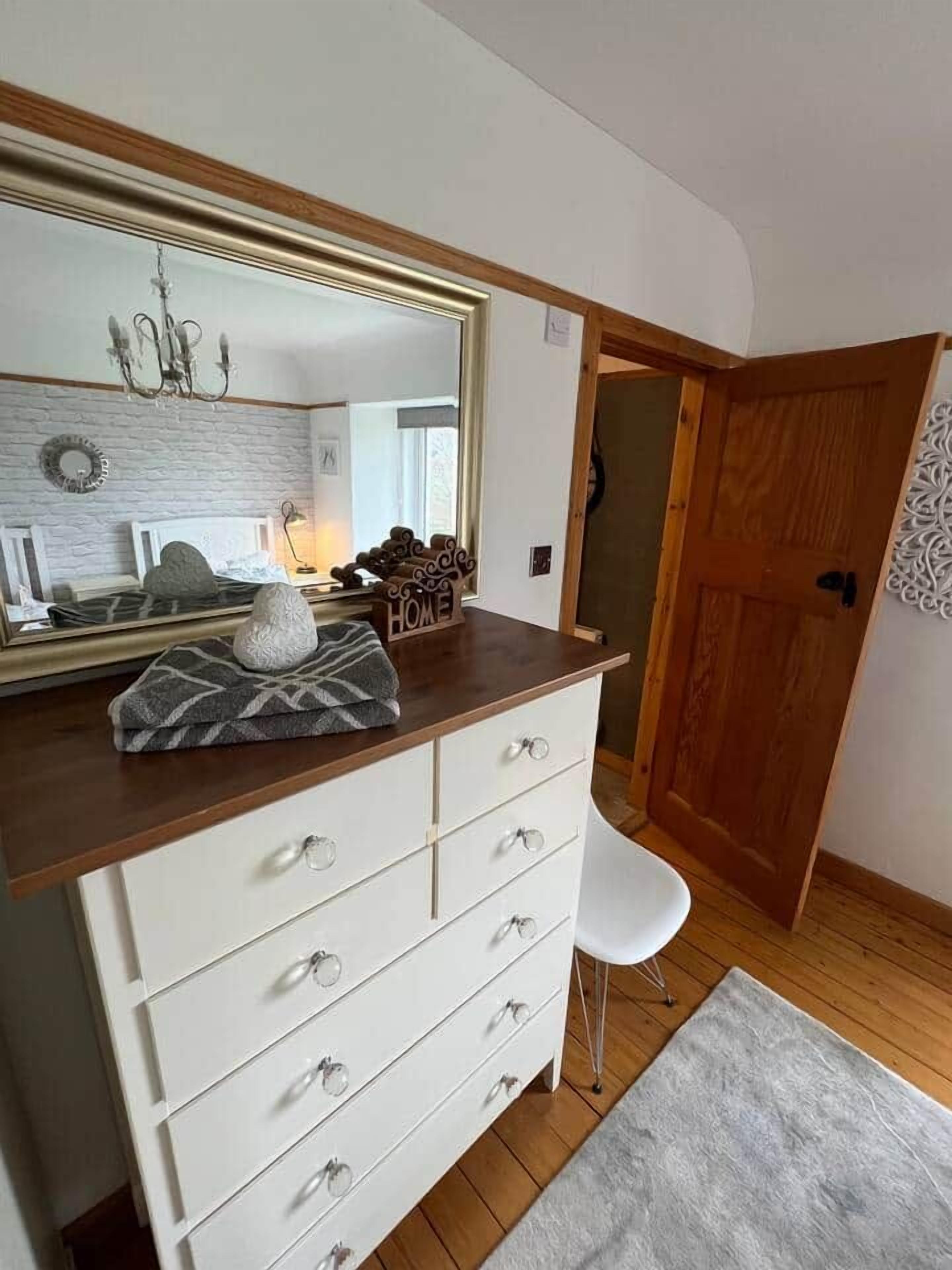 A bedroom with a large mirror, a dresser with drawers, a white chair, and a wooden door.