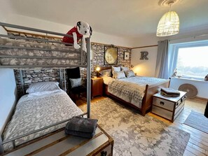 Une chambre avec des lits superposés, un mur en pierre, un lit recouvert d’une couvre-lit à motifs, une table de chevet avec une lampe et une fenêtre avec des rideaux.
