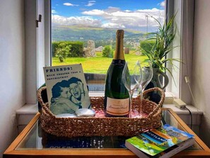 Una botella de champán, una copa, un libro y un cuadro en un aparador junto a una ventana con vistas al paisaje.