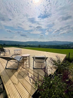 Una terraza de madera con mobiliario exterior y vistas a campos verdes y colinas lejanas.