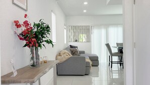 A modern living room with a grey sofa, a wooden side table with a vase of flowers, and a dining area with a table and chairs. - Private Yoga Space & Cold Plunge Near Everglades (Florida City)