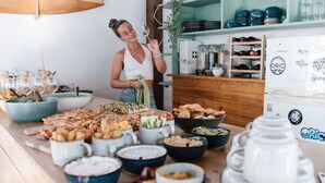 A buffet table with various dishes and drinks, a person serving, and kitchen appliances in the background. - Surfbase Lisbon (Almada)