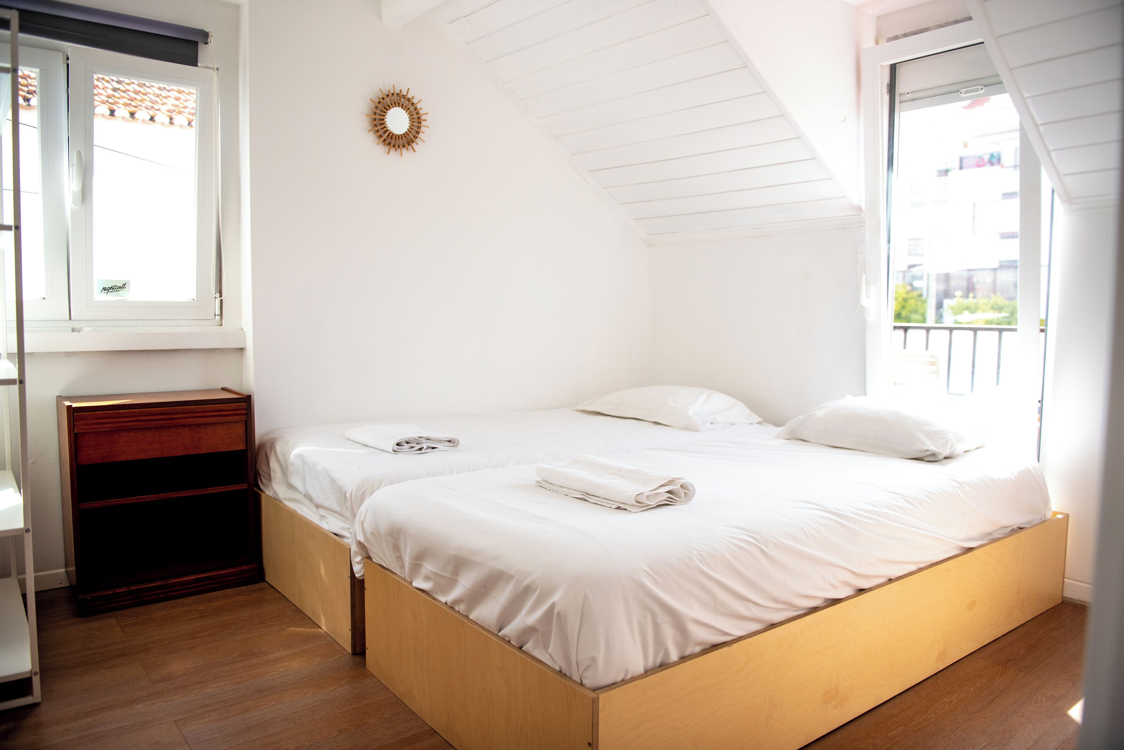 Un dormitorio con cama de madera, ropa de cama blanca, una mesita de noche de madera, una ventana con vistas y un adorno de pared con forma de sol.