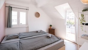 A bedroom with two beds, a window with curtains, a small table with a plant, and a view of the outside. - Surfbase Lisbon (Almada)