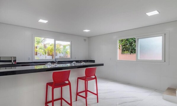 A modern kitchen with two red bar stools, a black countertop, and large windows offering views of greenery. - House in Condo w/ Pool Arraial D'ajuda Ort0003 (Porto Seguro)