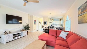Una sala de estar con un sofá rojo, un televisor montado en la pared, una zona de comedor con sillas, una cocina al fondo y un ventilador de techo.