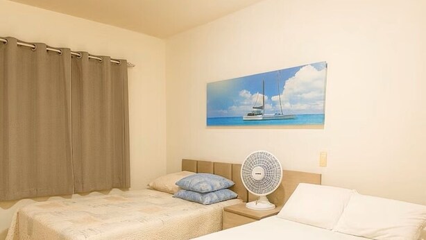 A hotel room with two beds, a bedside table with a fan, a chair, and a painting of a sailboat on the wall. - Apartment next to the Basilica of Saint Francis, Downtown Canindé, Ceará (Canindé)