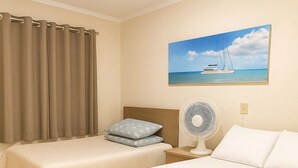 A hotel room with two single beds, a nightstand with a fan, a painting of a sailboat, and a curtain. - Apartment next to the Basilica of Saint Francis, Downtown Canindé, Ceará (Canindé)