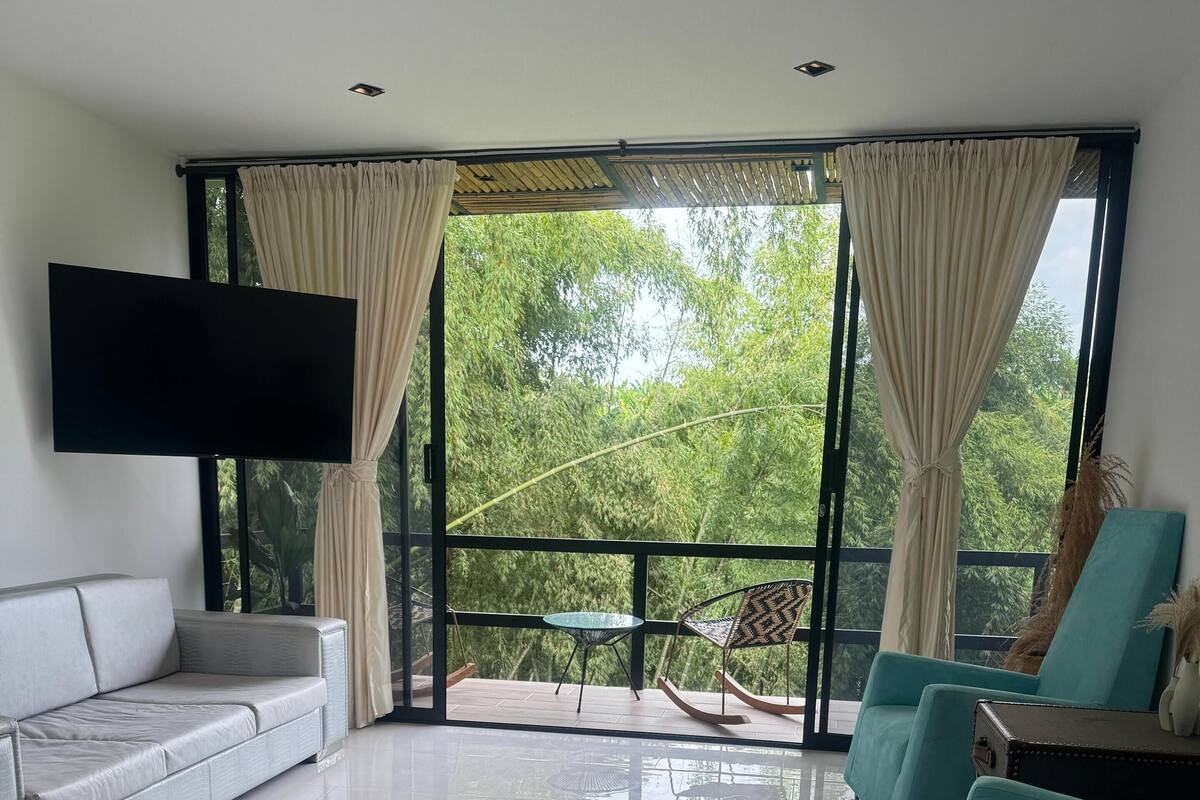 Une chambre d’hôtel moderne dotée d’un grand lit, d’un canapé, de deux fauteuils, d’une petite table et d’un téléviseur fixé au mur. La chambre dispose de grandes fenêtres avec des rideaux et offre une vue sur une végétation luxuriante.