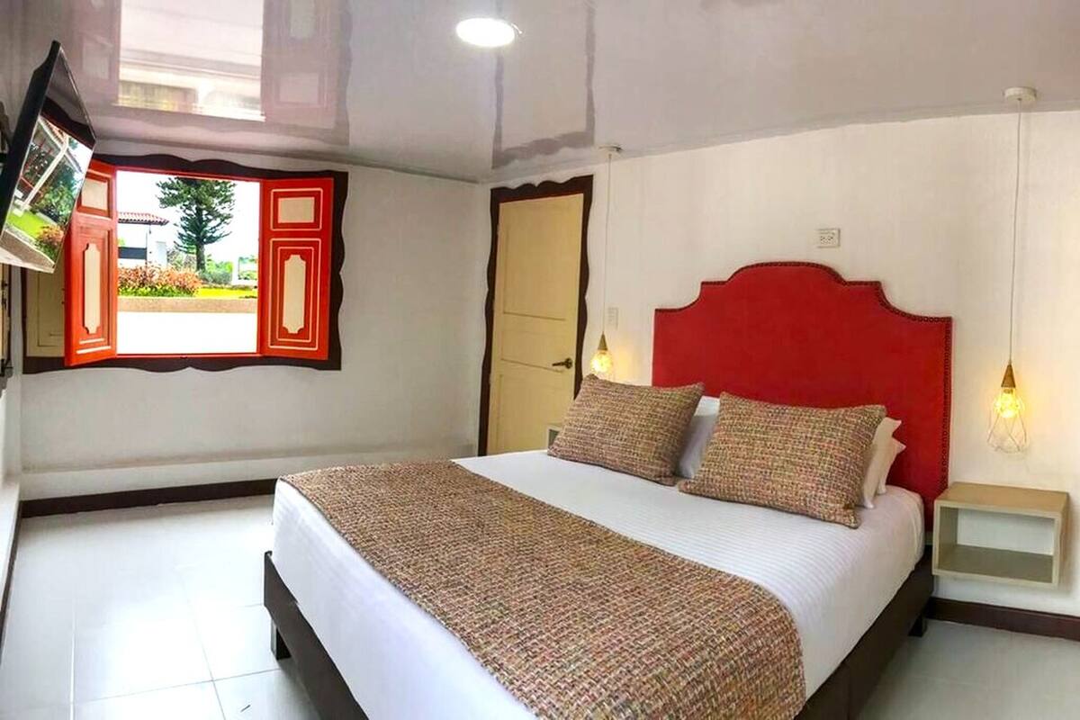 Une chambre d’hôtel avec un lit, une table de chevet, une tête de lit rouge et une fenêtre donnant sur un immeuble et de la verdure.