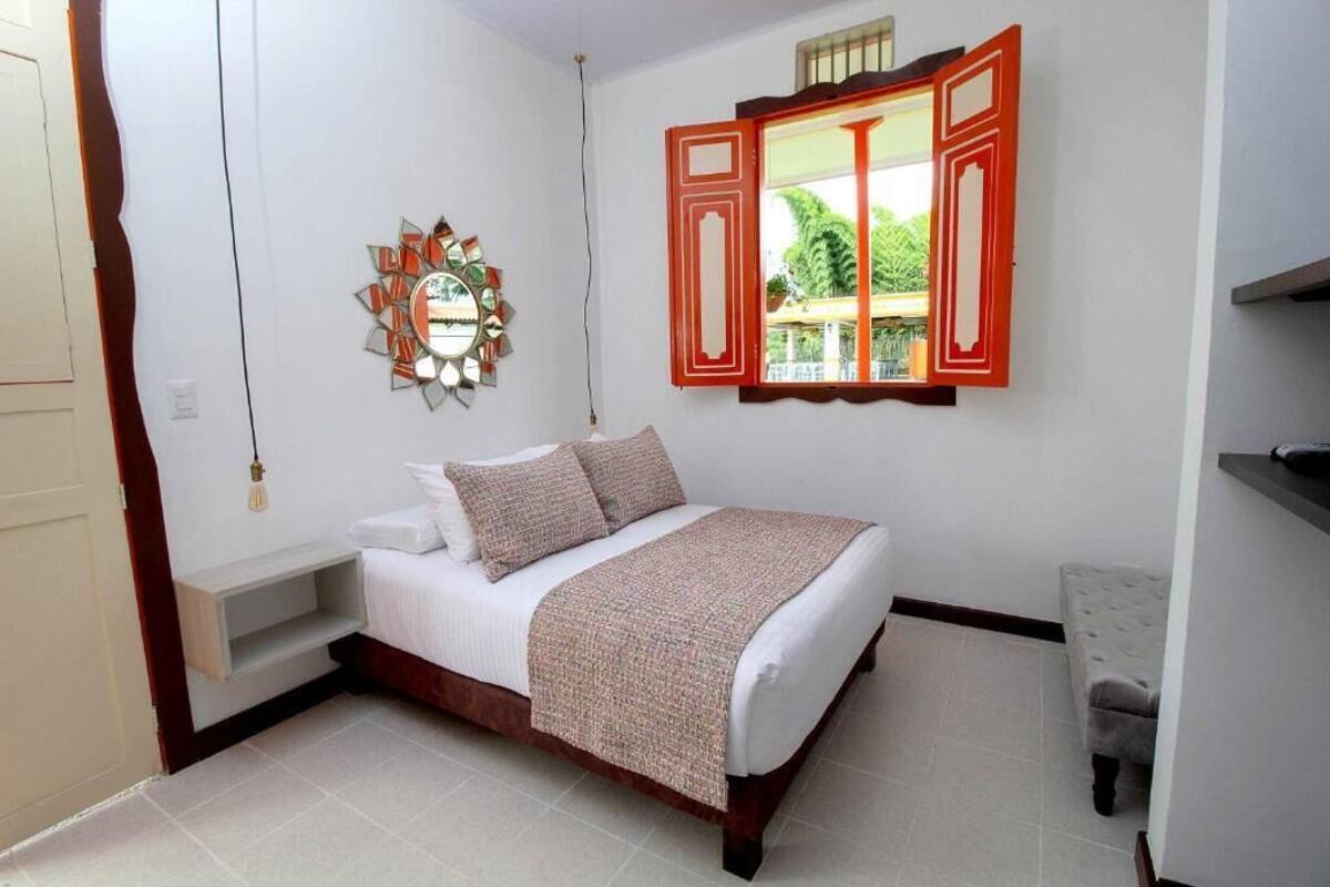 Une chambre à coucher avec un lit, un miroir fixé au mur et une fenêtre avec des volets rouges.