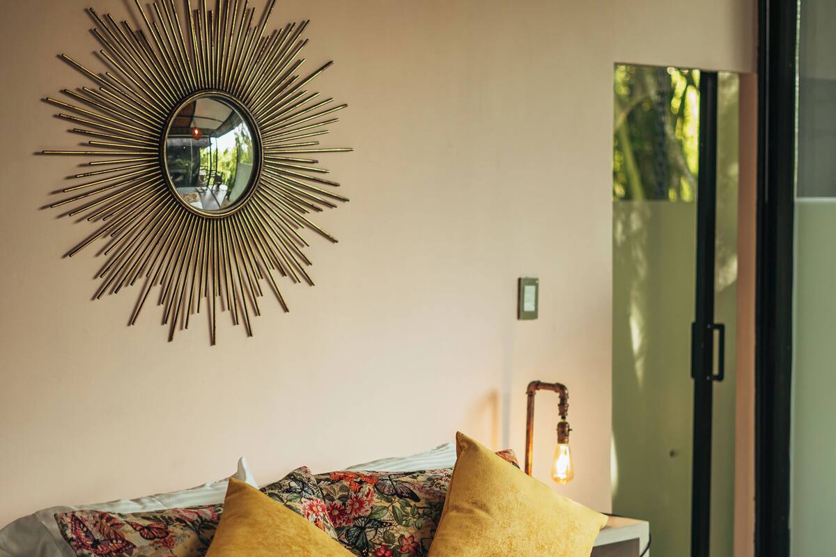Un lit avec une literie à motifs floraux et deux oreillers, un miroir fixé au mur et une lampe de chevet.