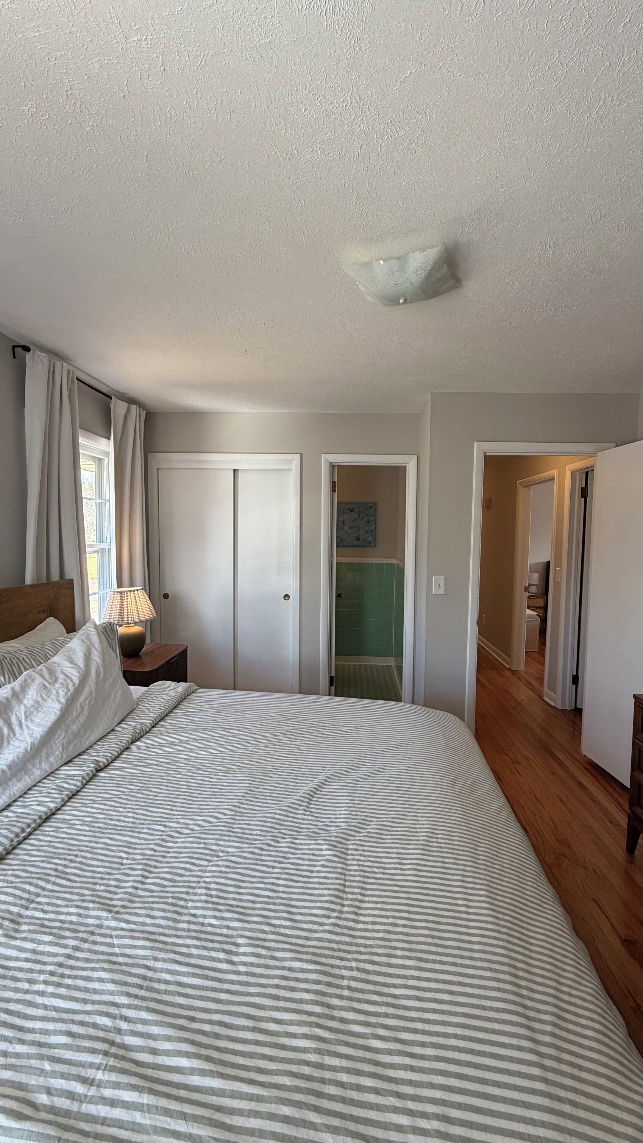 Un dormitorio con una cama grande, una mesita de noche con lámpara, una puerta que da al baño y un pasillo.