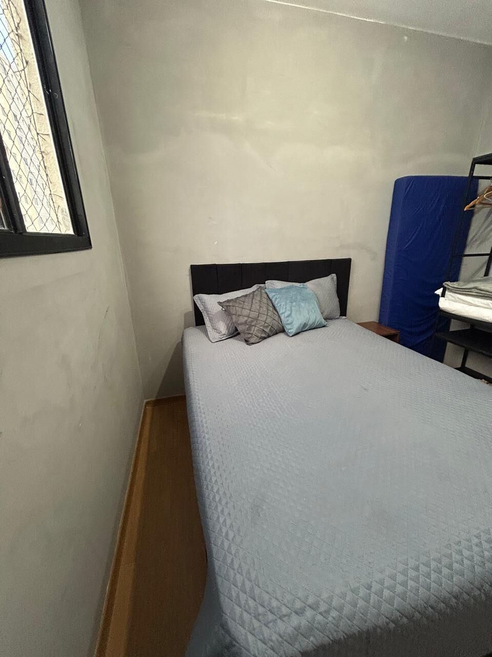 Une petite chambre avec un lit simple, un mur uni et une fenêtre munie d’un grillage métallique.