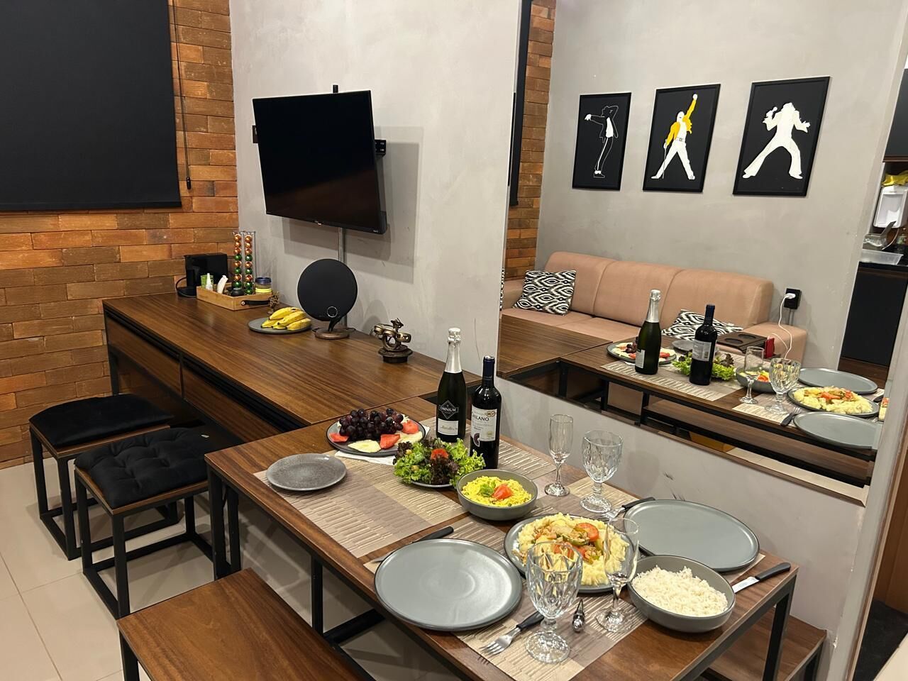 A modern dining area with a long table set for a meal, a TV mounted on the wall, and a sofa in the background.