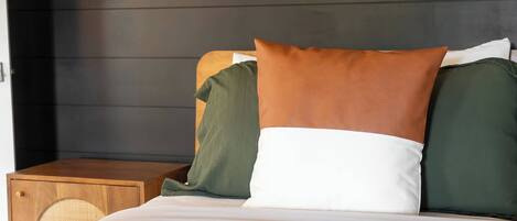 Una cama con colcha blanca y verde, una mesita de noche de madera y una silla de mimbre, junto a un cabecero de madera oscura.