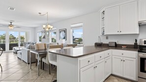 A spacious kitchen with white cabinetry, a dining area with a table and chairs, and a view through the glass doors. - Sandy Pelican by Avantstay Exclusive Beach Access + Private Pool + Firepit + Balcony Sunset Views (Panama City Beach)