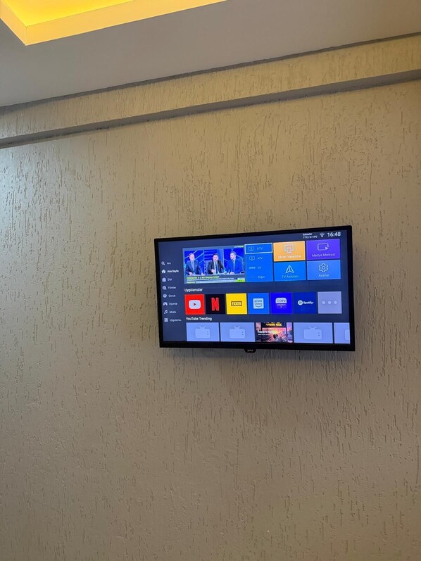 Un téléviseur à écran plat fixé à un mur texturé affiche une interface de guide de programmes télévisés, avec différentes plateformes de diffusion en continu et une section présentant les vidéos les plus populaires sur YouTube.