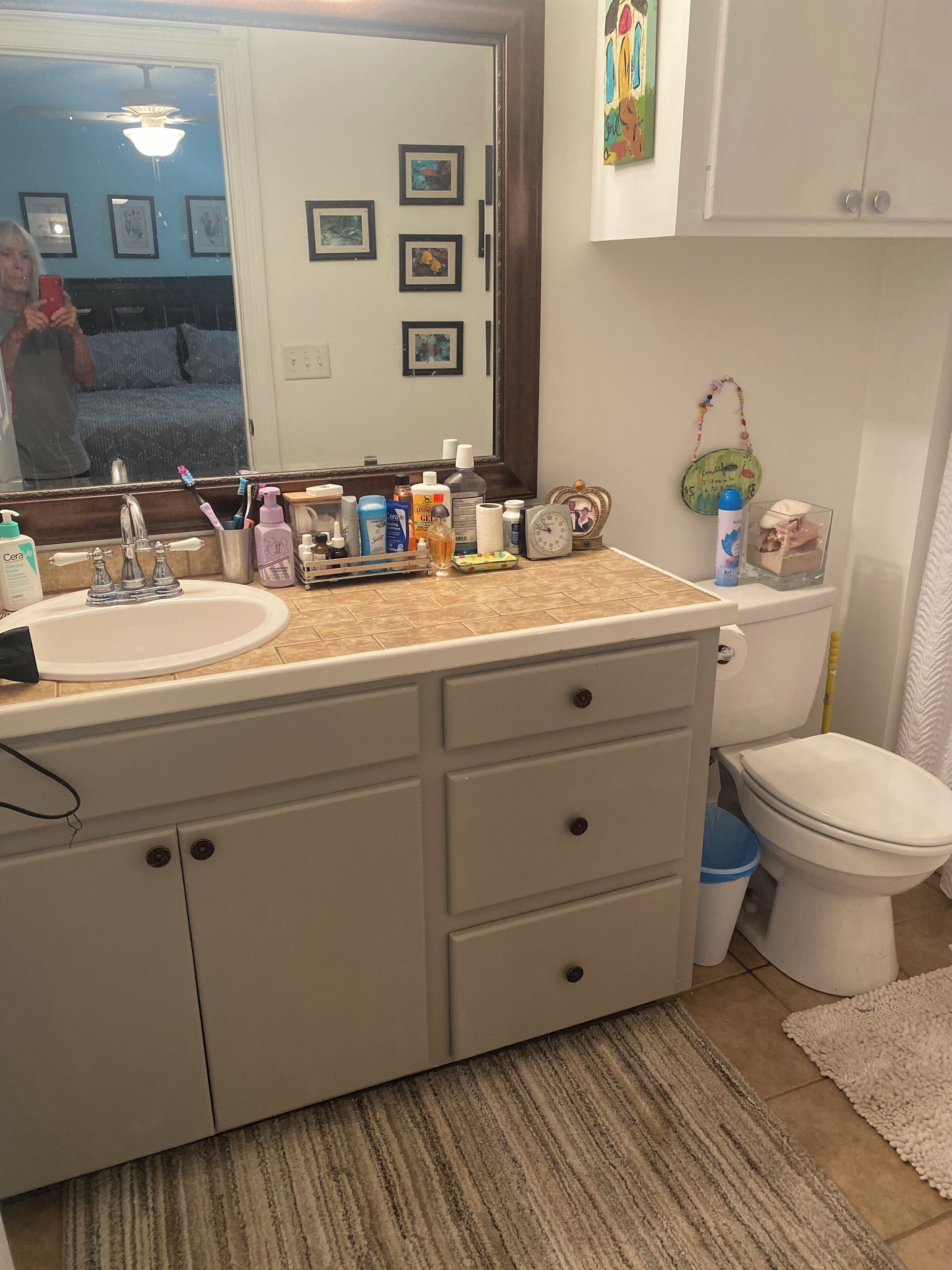 A bathroom with a sink, mirror, and toilet. There are toiletries on the sink counter, a clock, and a framed picture on the wall.