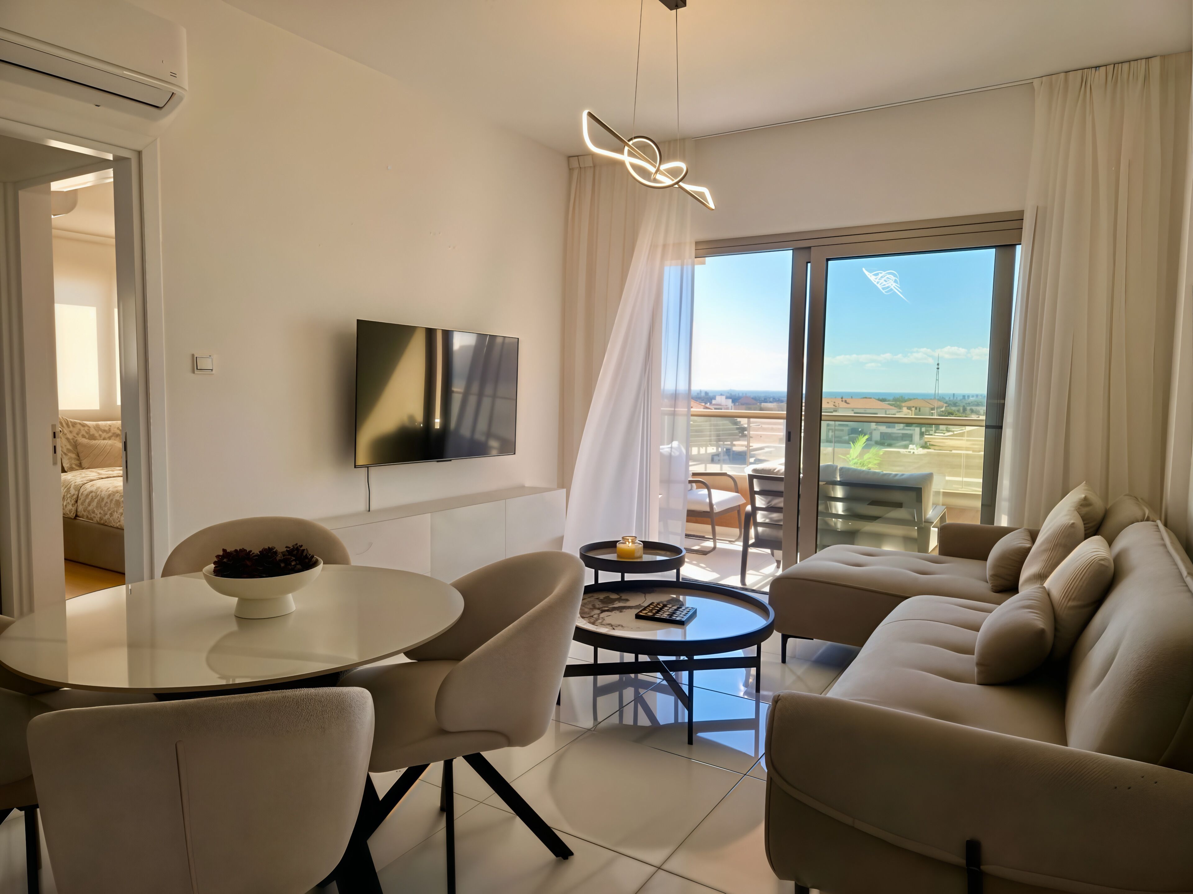 A modern living room with a flat-screen TV, a round dining table with chairs, a sofa, and a balcony with a view.