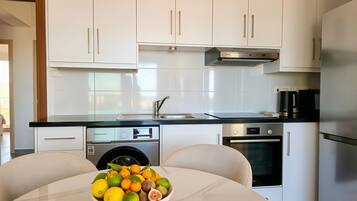 A modern kitchen with white cabinets, a stainless steel range hood, a built-in oven, and a bowl of fruit on the table.
