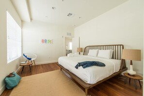 Ein Schlafzimmer mit einem hölzernen Bett, weißer Bettwäsche, einem blauen Sessel, einem kleinen Tisch und einem Fenster mit Jalousien.