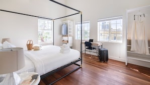 Une chambre d’hôtel moderne avec un lit à baldaquin, un bureau avec un ordinateur portable, une chaise et une valise. Des fenêtres laissent entrer la lumière naturelle et offrent une vue sur la ville.