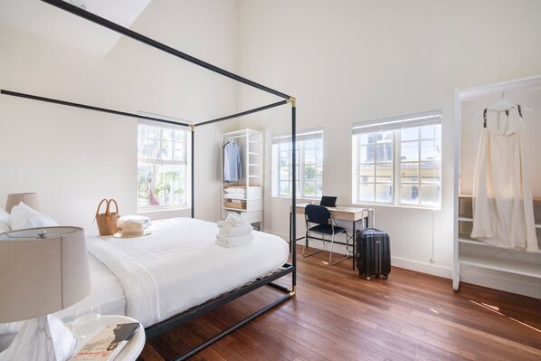 A modern hotel room with a four-poster bed, a desk with a laptop, a chair, and a suitcase. There are windows providing natural light and a view of the city. - 4-bedroom Gem Sleeps 10 Best Value in Sobe (Miami Beach)