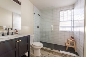 Ein Badezimmer mit Toilette, dunklem Holzwaschtisch, einer Duschkabine aus Glas und einem kleinen Holzschemel.