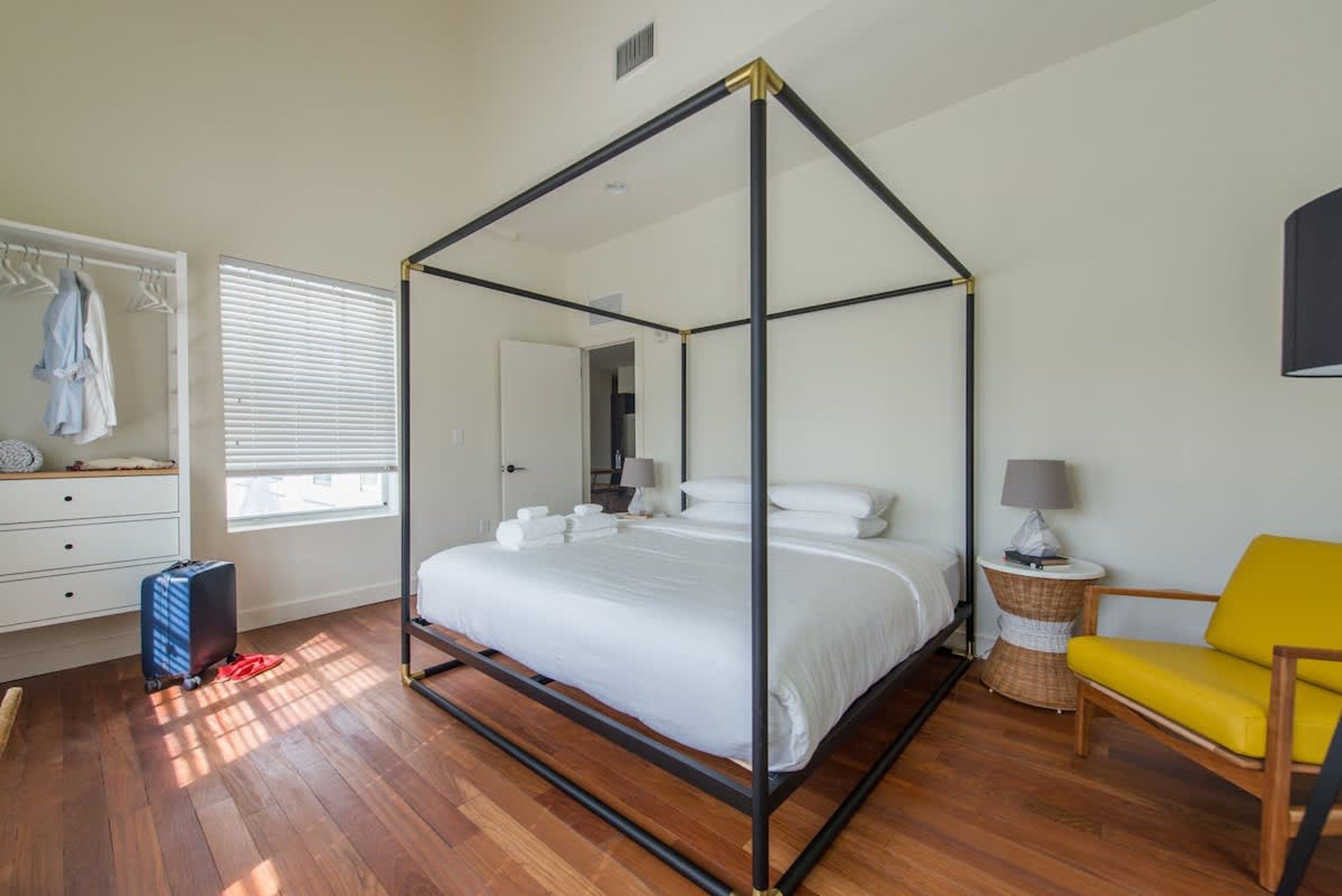 Una cama con dosel con sábanas blancas, una maleta, un sillón amarillo y una mesita redonda con una lámpara.
