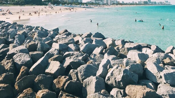 Une plage avec de gros rochers au premier plan, une étendue de sable et des bâtiments en arrière-plan.