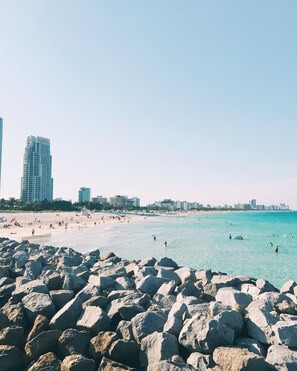 Una playa con grandes rocas al frente, una orilla de arena y edificios en la distancia.