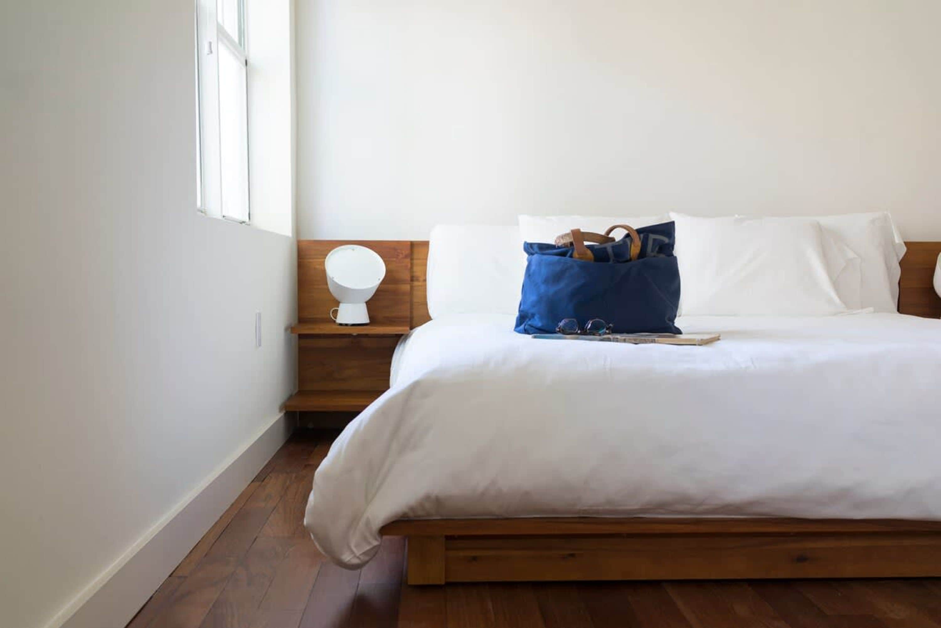 Une chambre à coucher avec un lit en bois, du linge de lit blanc, une table de chevet avec une lampe et une fenêtre.