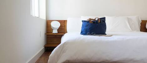 Une chambre à coucher avec un lit en bois, du linge de lit blanc, une table de chevet avec une lampe et une fenêtre.