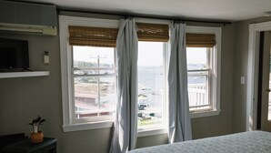 Une chambre à coucher avec un lit, un téléviseur posé sur une étagère et une vue sur la mer à travers deux fenêtres équipées de rideaux.