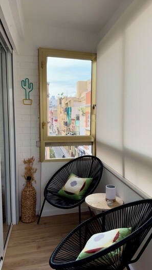 Ein Balkon mit zwei schwarzen Drahtstühlen, einem kleinen runden Tisch mit einer weißen Tasse und Blick auf bunte Häuser.