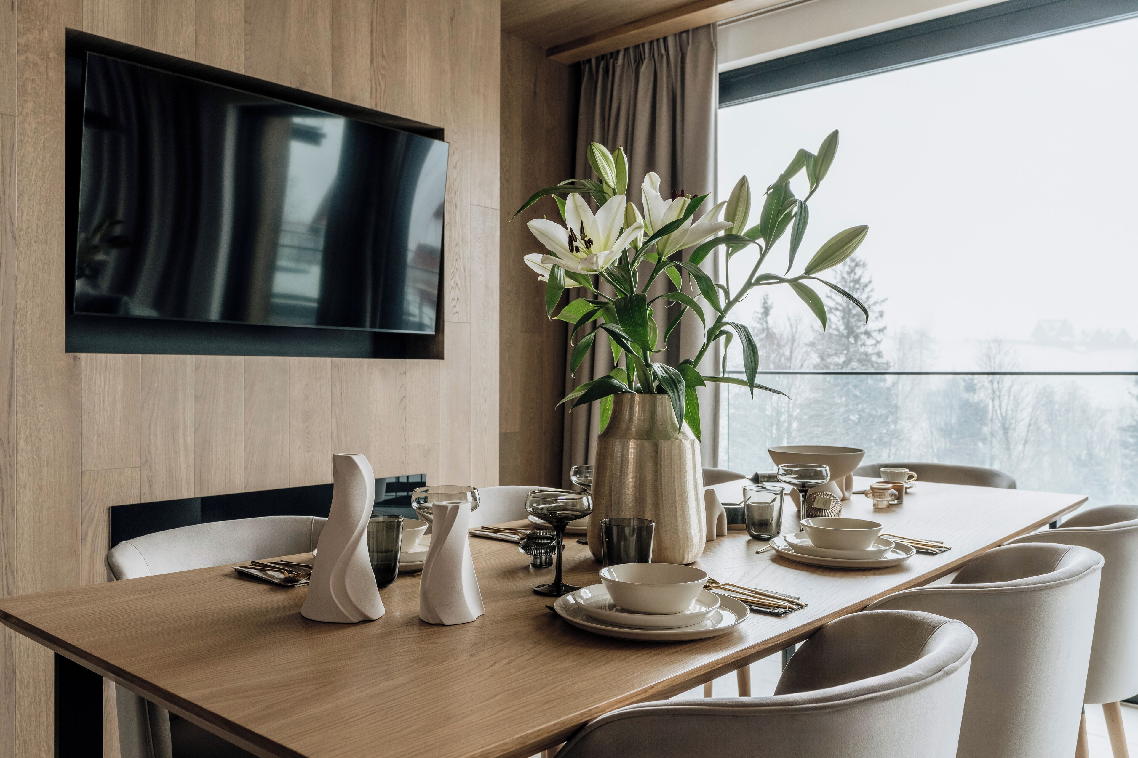 Un comedor con mesa de madera, sillas blancas, un jarrón con flores y un televisor montado en la pared.