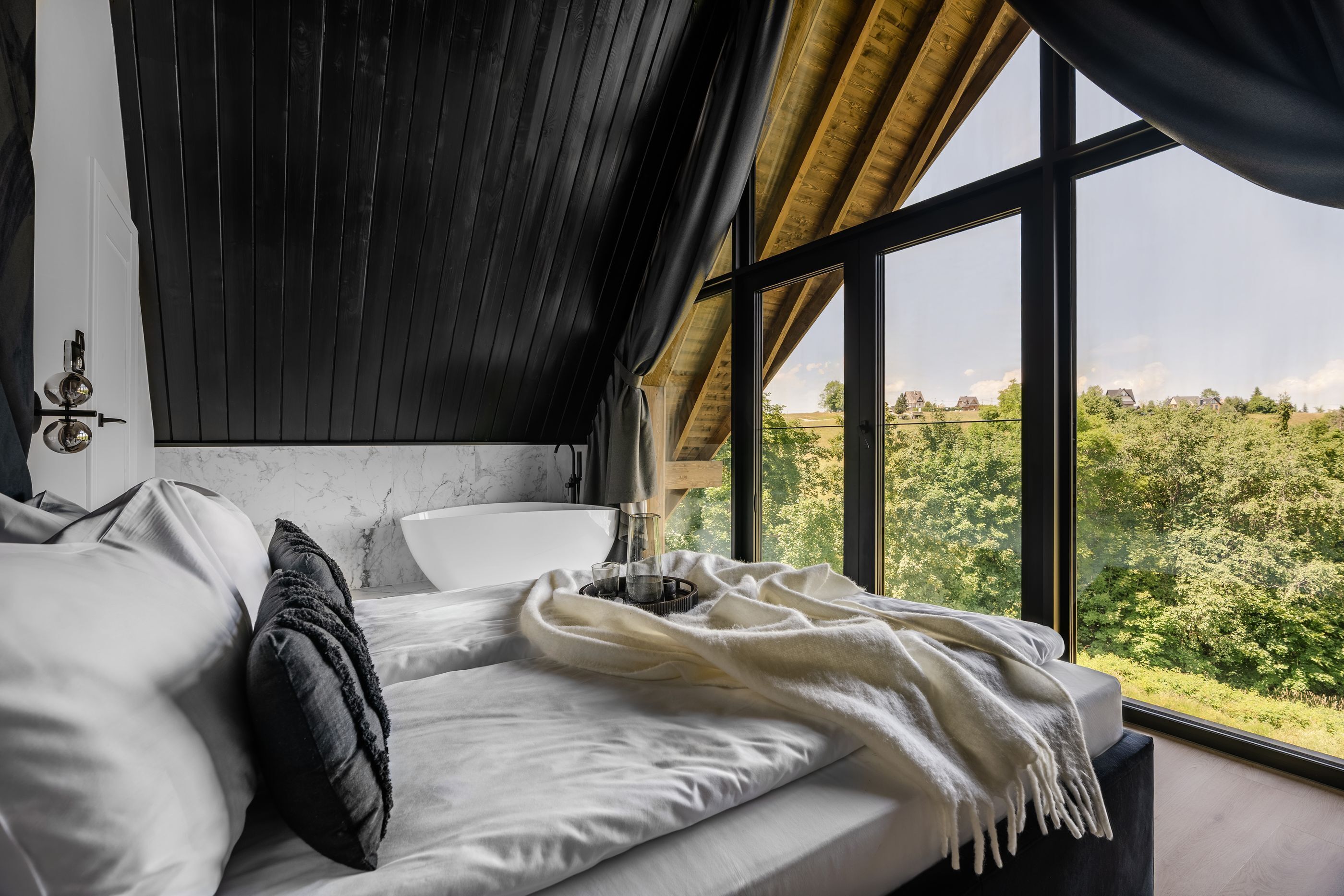 A bedroom with a large window, a bed with white bedding, a dark ceiling, and a view of greenery outside.
