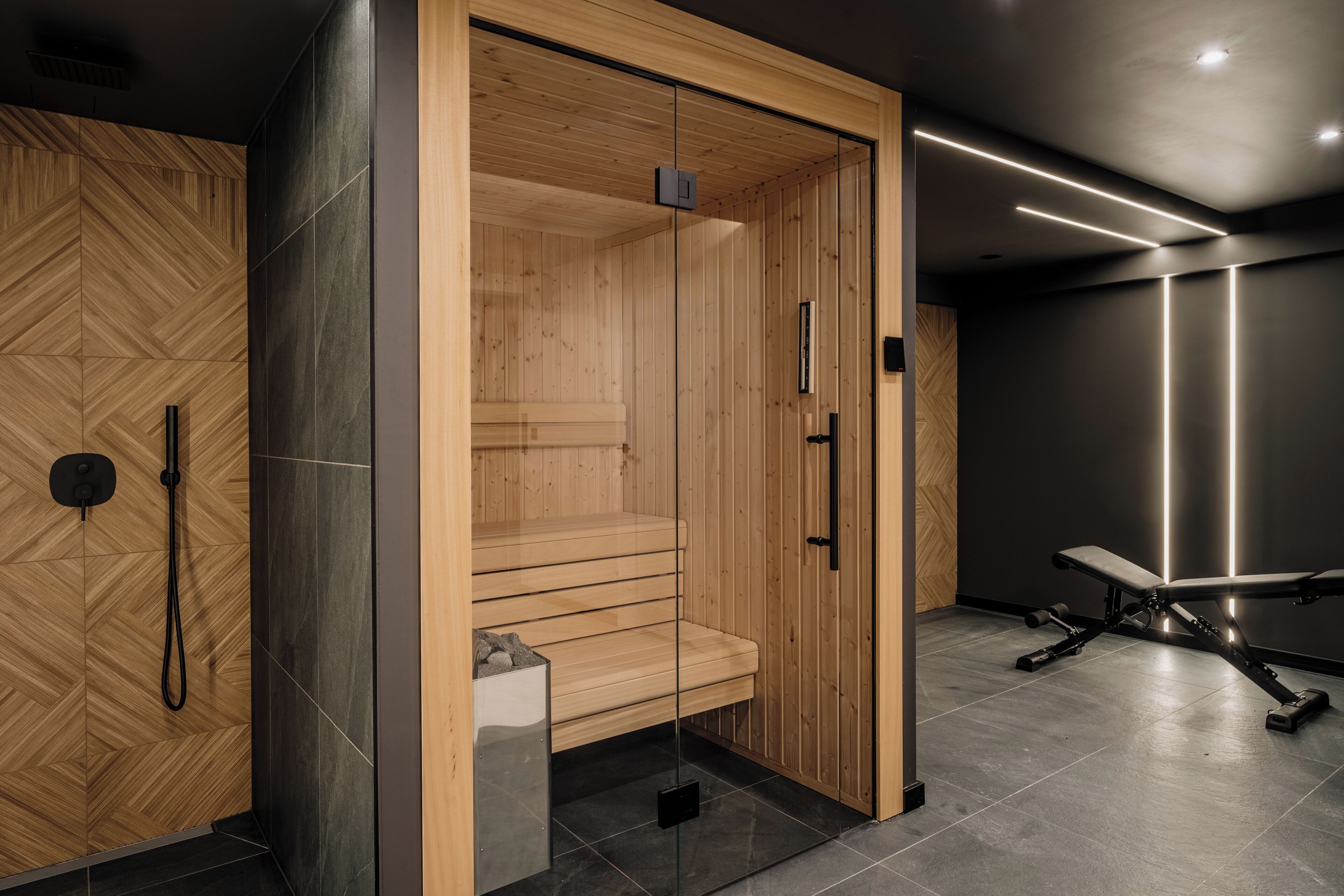 A modern sauna with wooden walls and a bench, a glass door, and a bench with weights in the adjacent room.