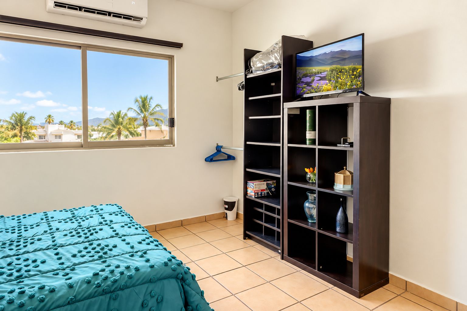 Een slaapkamer met een bed, een tv op een standaard, een raam met uitzicht en een plank met diverse spullen.