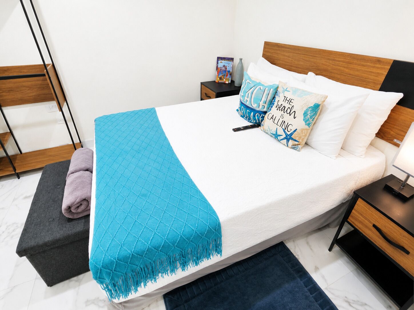 A neatly made bed with a blue blanket, white pillows, and a beach-themed pillow. There is a nightstand with a lamp and a book.