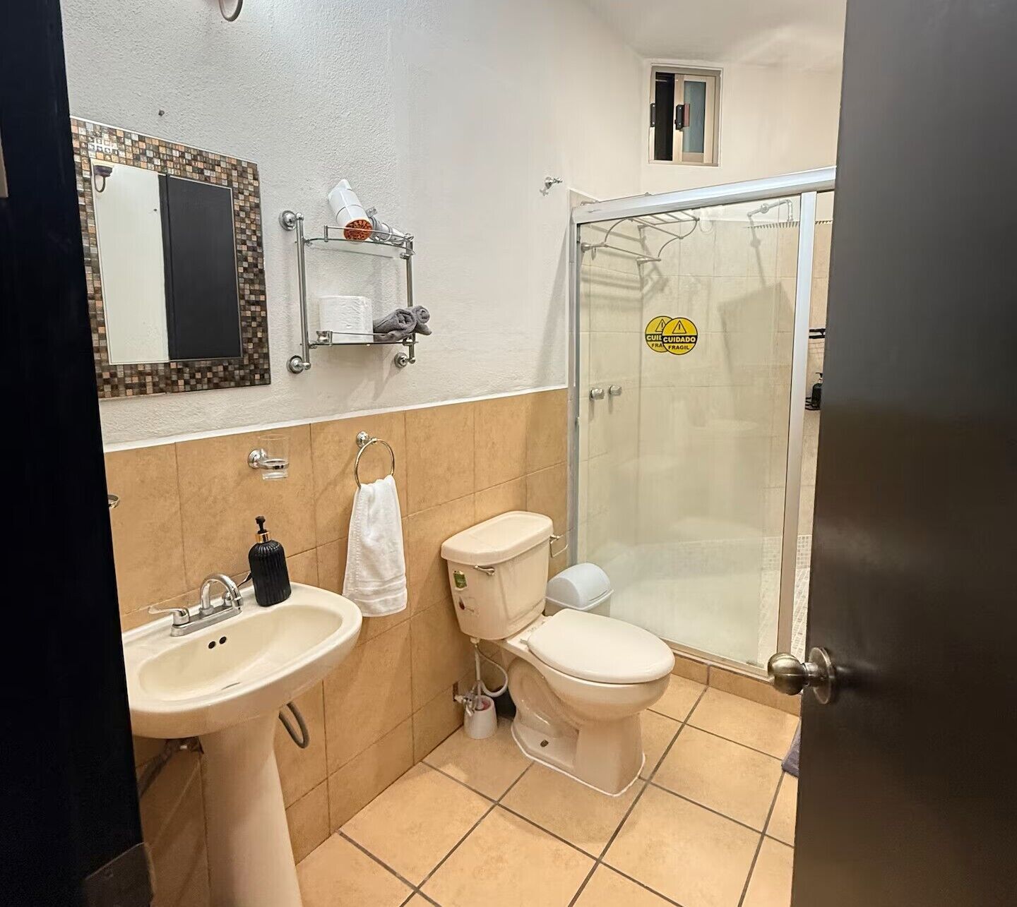 Un baño con lavamanos, inodoro y ducha. Hay un perchero y una pequeña ventana sobre el inodoro.