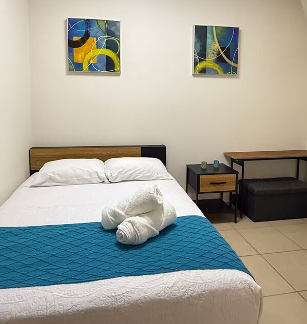 Ein Hotelzimmer mit einem Bett, zwei abstrakten Bildern an der Wand, einem Nachttisch, einer Schublade und einer Bank.