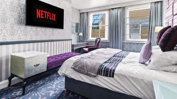 Quarto de hotel moderno com uma cama grande, televisão na parede, escrivaninha e janela com cortinas.