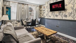 Uma sala de estar com sofá, mesa de centro de madeira, área de jantar com cadeiras e mesa, e uma televisão na parede exibindo 'NETFLIX'.