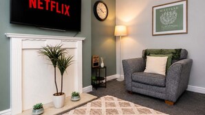 A living room with a fireplace, a Netflix TV, a clock, a lamp, a framed artwork, and a grey armchair.