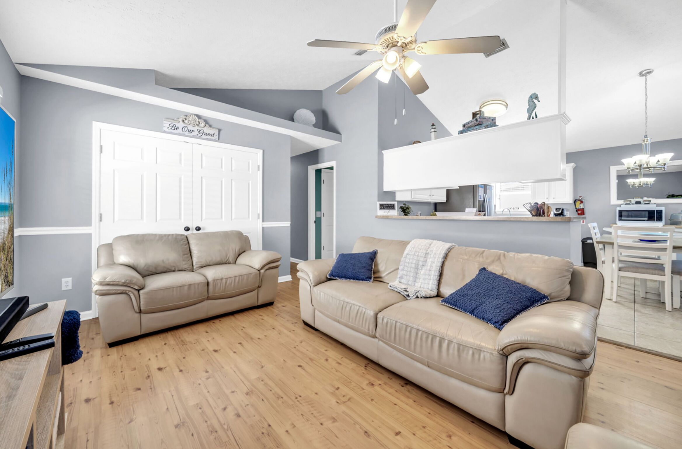 A modern living room with a beige sofa, a ceiling fan, and a kitchen area with white cabinets and a dining table.