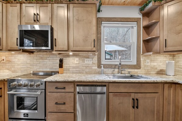 A kitchen with wooden cabinets, a stainless steel microwave, a built-in oven, a dishwasher, and a window with a view of trees.