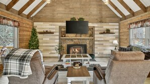 A cozy living room with a fireplace, wooden beams, a television, and a Christmas tree.