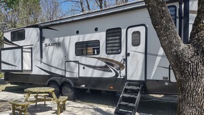 Un camper bianco con porta scorrevole e un piccolo oblò, parcheggiato su una superficie in cemento con un tavolo da picnic e una scala nelle vicinanze.